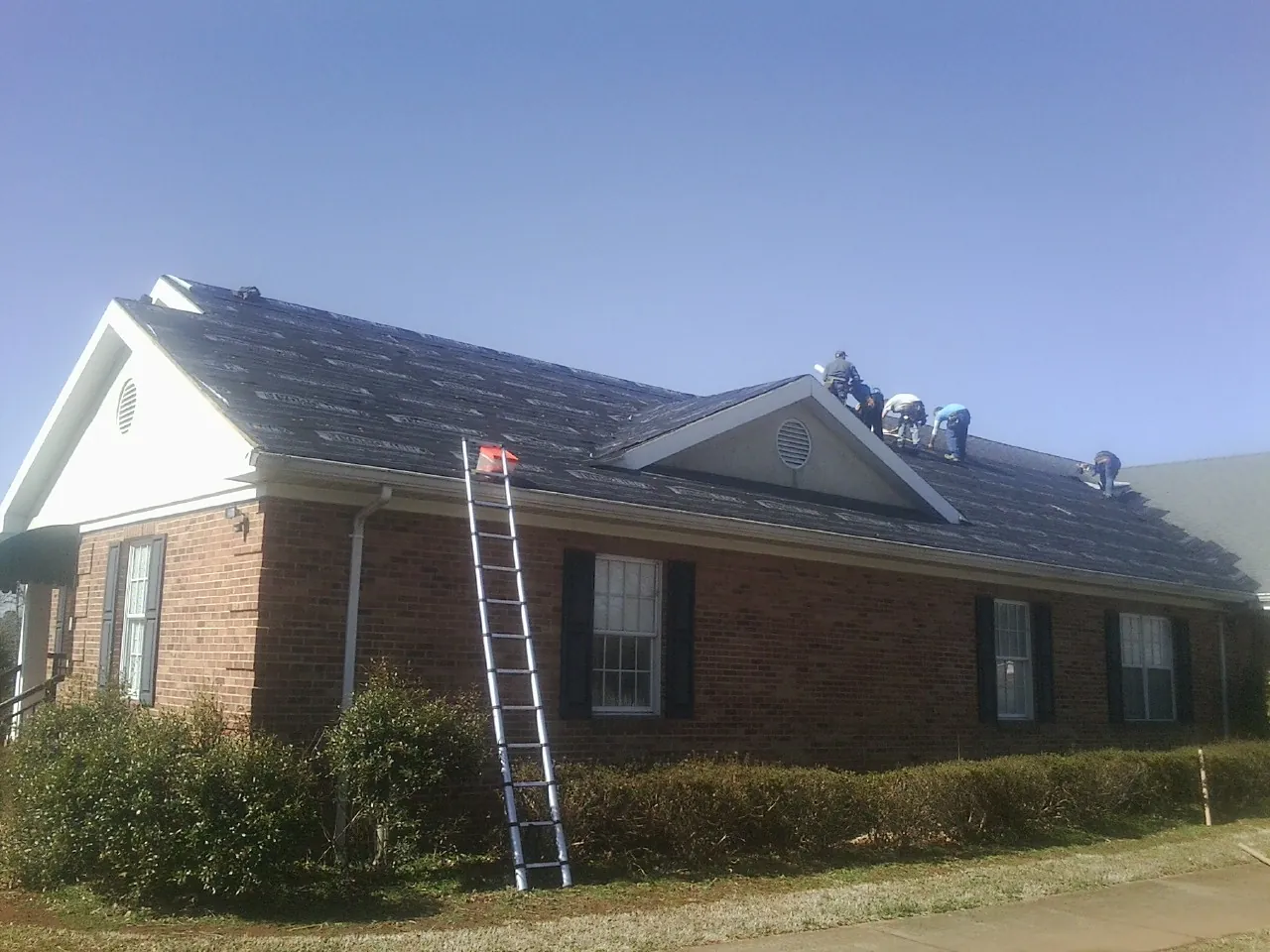 Crew tearing shingles off a brick ranch roof with a ladder set — Metal Roof Repair work by our crew in Newton Grove