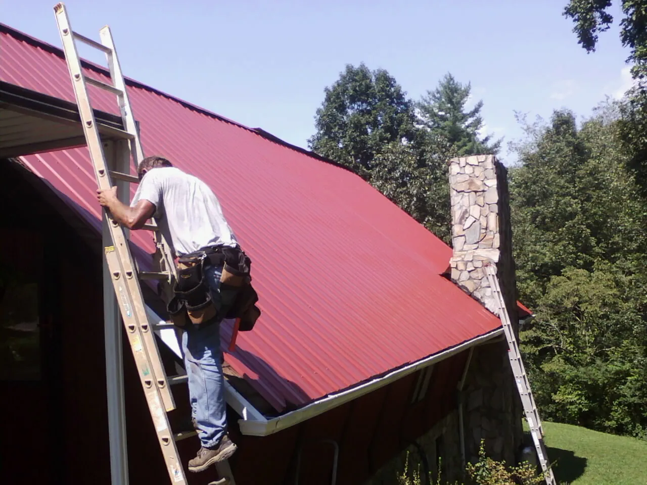 Roofer on a ladder beside a red metal roof and stone chimney — Soffit & Fascia Repair by Roofing Brothers in Newton Grove
